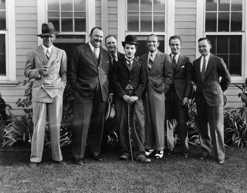 Charlie Chaplin, dressed as The Tramp, standing outdoors with Fairbanks Jr, bandleader Paul Whiteman, and with Fairbanks Sr third from the right.