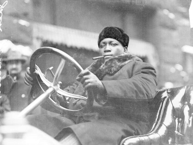 1910, Jack Johnson smoking a cigar while driving an automobile in Chicago, Illinois. Photo by Chicago History Museum.