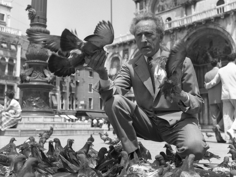 Jean Cocteau wearing a plaid suit and a fancy tie, crouched in St. Mark Square while feeding pigeons, Venice 1956. Photo by Archivio Cameraphoto Epoche/Getty Images.