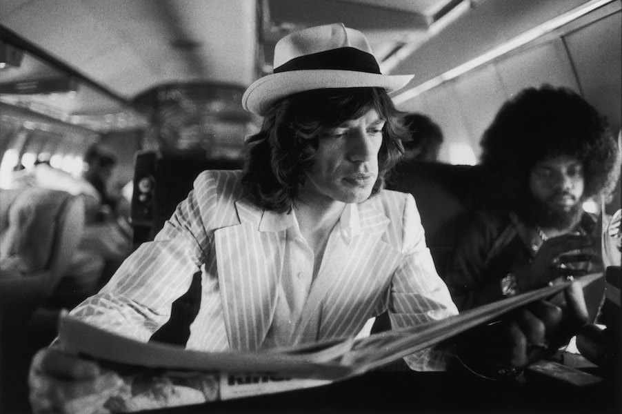 Mick Jagger keeps abreast of current affairs while travelling between concerts in the Rolling Stones' private jet during their 1975 Tour of the Americas. Also aboard is American keyboard player Billy Preston (right). Photo by Christopher Simon Sykes/Hulton Archive/Getty Images.