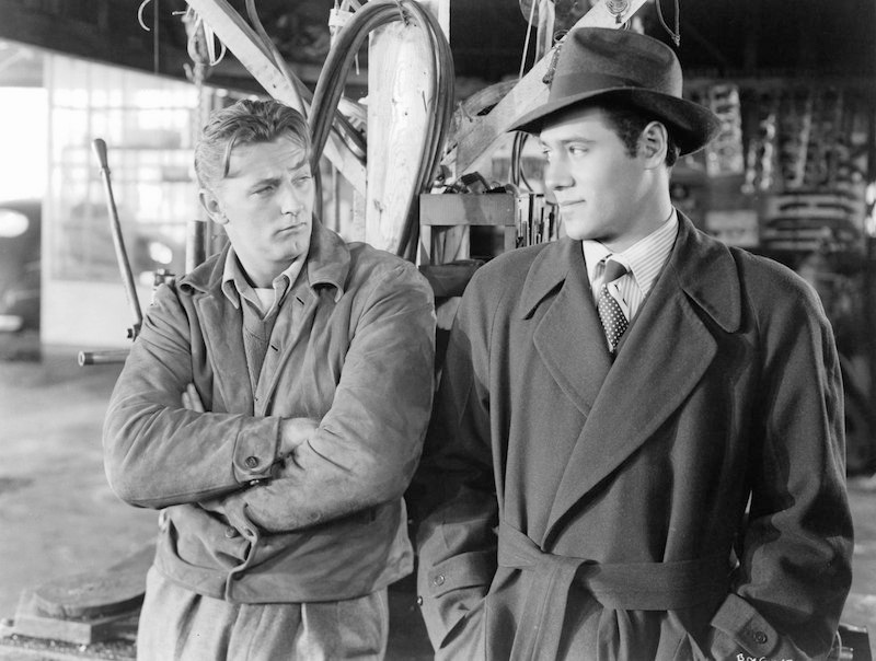 Robert Mitchum with arms folded looking over at Steve Brodie in a scene from the film Out Of The Past, 1947. Photo by RKO Radio Pictures/Getty Images.