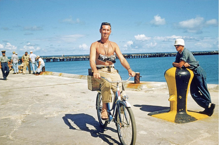 Slim Aarons riding a bicycle with his cameras around his neck, 1955.