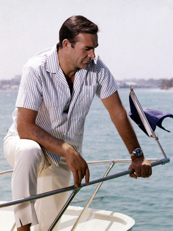 Connery’s Bond wearing a Bengal-striped, camp collar shirt with white trousers that feature a distinct slit at the hem. Notice also Bond’s Breitling Top Time wristwatch, which famously featured a geiger counter, as fitted by Q Branch. Connery is pictured on the set of Thunderball in Nassau, the Bahamas in 1965.