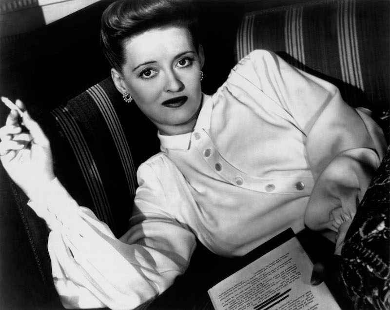 Elegantly reading whilst smoking a cigarette on the set of Now Voyager, 1942.