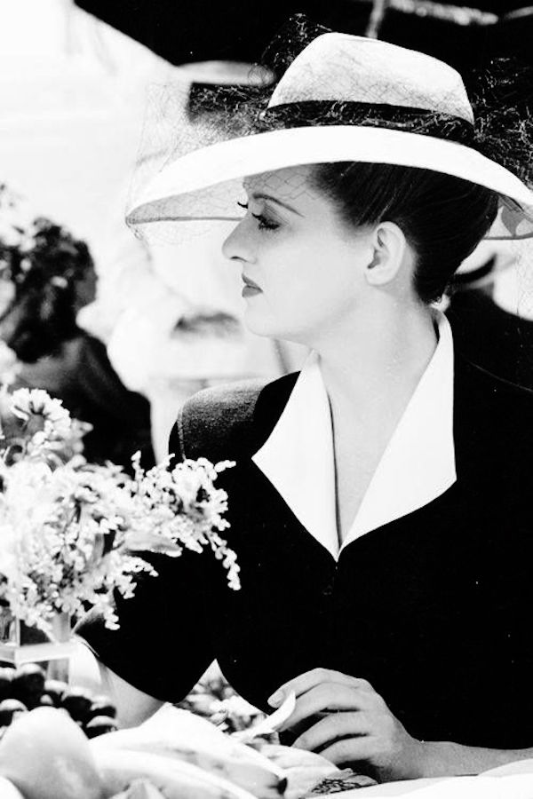 Wearing a semi-veiled straw hat, open collar shirt and short-sleeved blouse, circa 1940s.