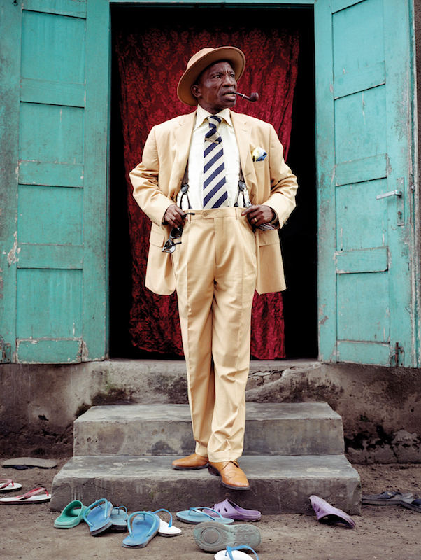 Severin Mouyengo, who has been a sapeur since the seventies, poses in the entrance of his family house in the Bacongo neighborhood. Sandals on the ground are from his family members. In the Congo, as in other parts of Africa, people often take their shoes off before entering home.