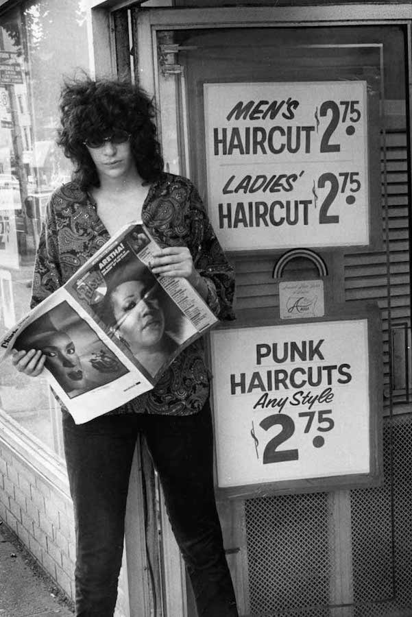 Joey Ramone stands outside a hairdresser that specialises in ‘Punk Haircuts’ reading a copy of Long Island Pulse magazine with Aretha Franklin starring on the cover, circa 1968.