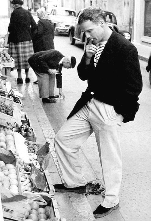 Buying fruit in the south of France, 1957. He wears wide-legged trousers, a boxy-cut blazer and a striped crew neck.