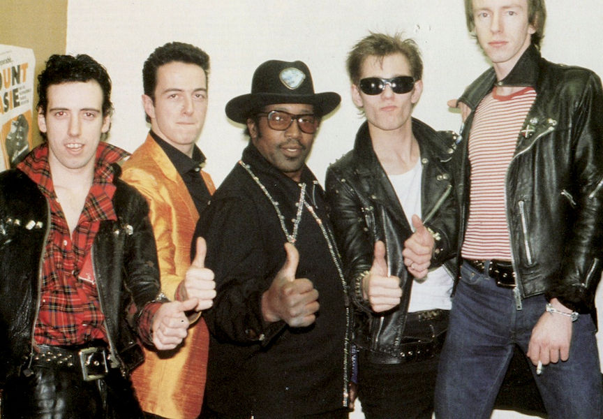 The Clash and Bo Diddley on tour in 1979. Left, Mick Jones sports a dishevelled tartan shirt with the collar turned up, tucked into a chunky leather belt and leather trousers.