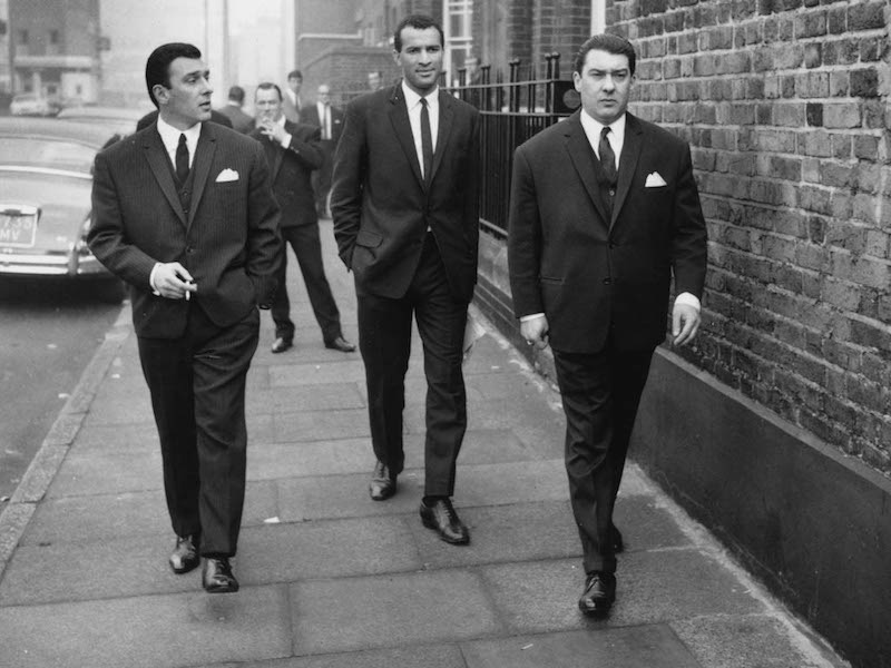 London gangsters Ronnie and Reggie Kray stroll along an East End street in matching single-breasted suits in London, 1965. Photo by Ron Gerelli/Express/Getty Images.