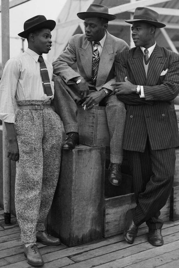 John Hazel, Harold Wilmot and John Richards arrive in Tilbury from Jamaica aboard the 'Empire Windrush', in 1948. Note the wide-legged, Hollywood top trousers on the right, which are cinched tightly with a slim belt. The pinstripe suit on the right is reminiscent of a zoot suit, albeit with less exaggerated proportions.