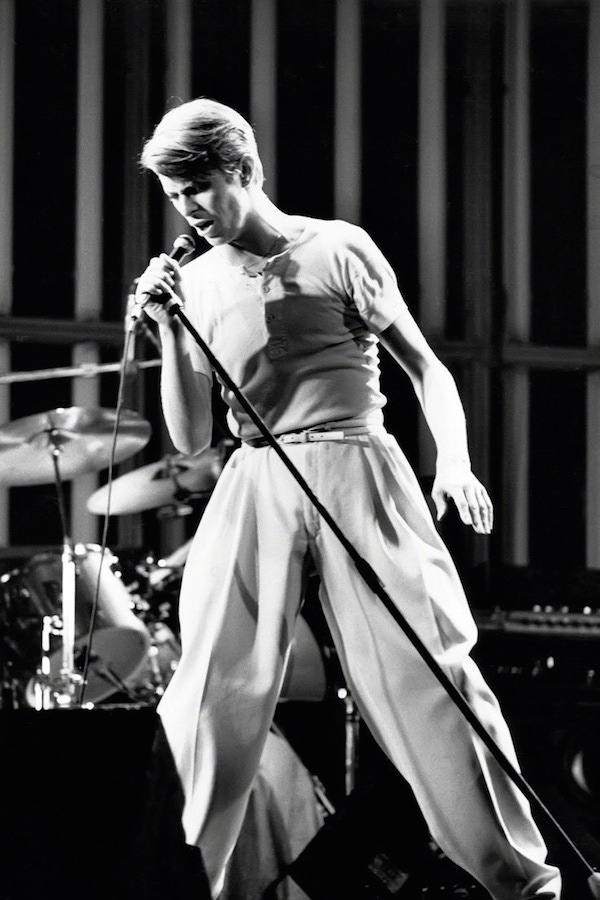 David Bowie displaying his wide trouser prowess, as he caresses the mic, circa 1970s. Note the skinny belt, which accentuates the high-waist and plush swathes of fabric.