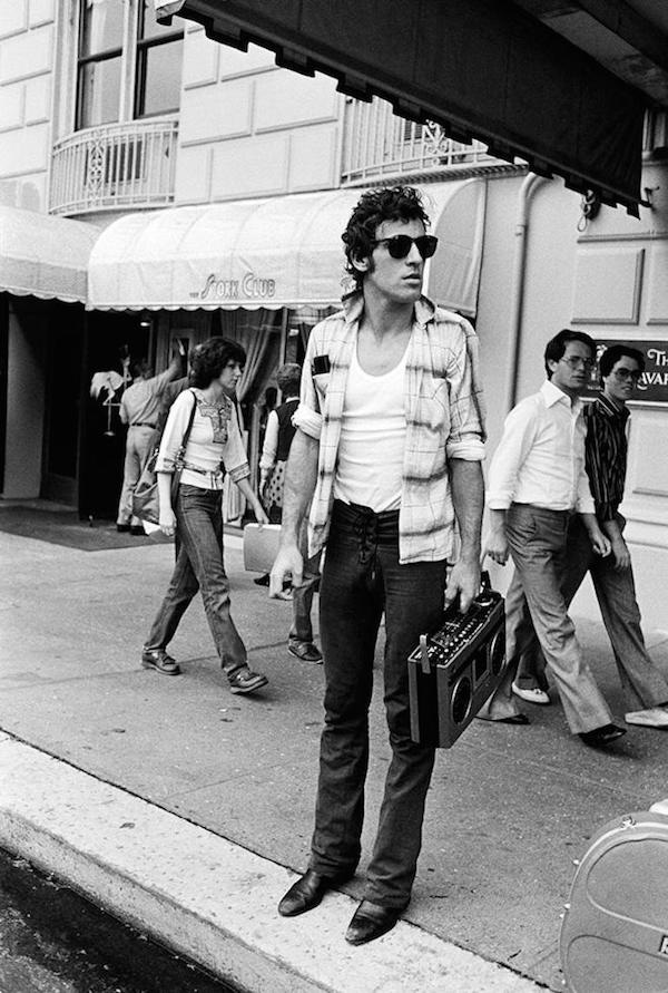 Holding a boombox on a New York sidewalk whilst wearing an open checked shirt on top of a white T-shirt and jeans paired with sunglasses, 1978. Photograph by Lynn Goldsmith.