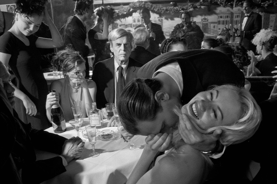 Writer Jared Stern kisses a lady friend as George Plimpton looks on while dining with friends at Elaine's, 1999. Visible in the background is Steve Garbarino, Ivanka Trump and a reflection of the photographer Larry Fink. Photo by Larry Fink/Courtesy Aperture.