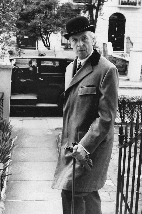 English photographer Cecil Beaton wears a low Derby hat and covert coat with flapped breast pockets, 1955.