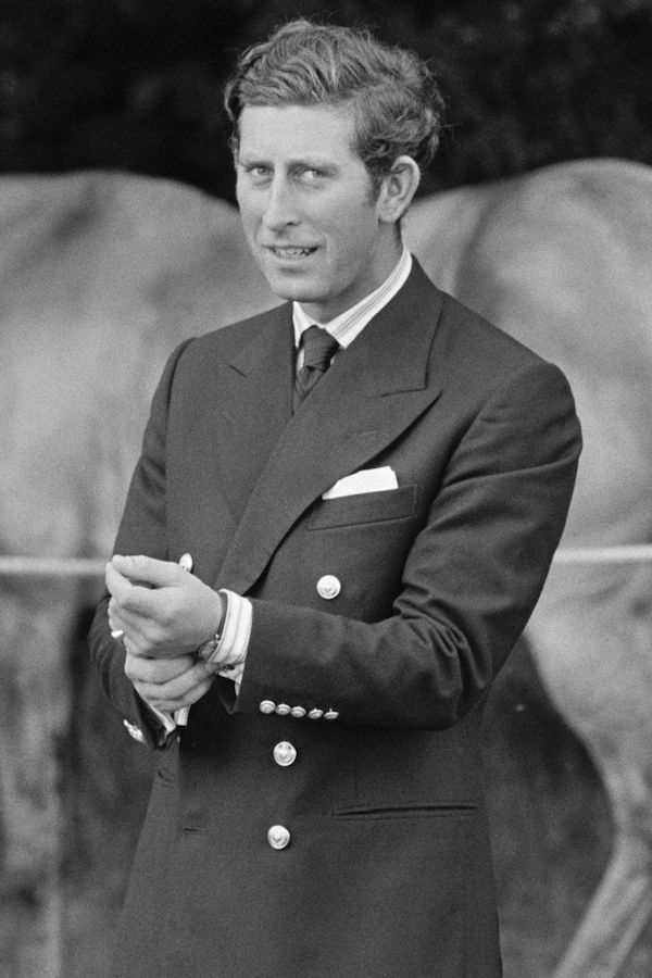 A young Prince Charles adjusts his sleeve, wearing a double-breasted navy blazer with peaked lapels and decorative gold buttons at the cuff, finished with a tie and neatly folded handkerchief in his outbreast pocket.