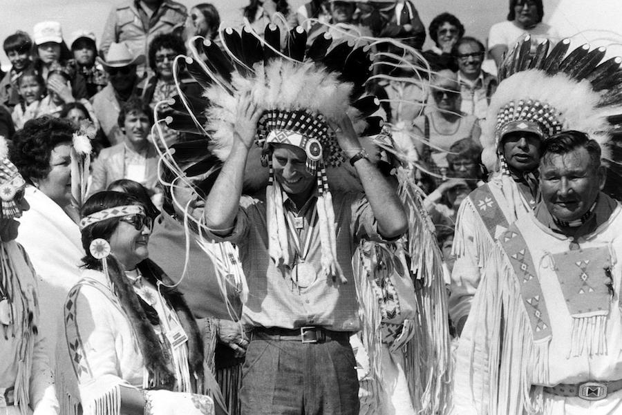 Prince Charles attending a Blackfoot Indian ceremony in Calgary, Canada, 1977, wearing a tribal headdress and short-sleeved linen shirt.