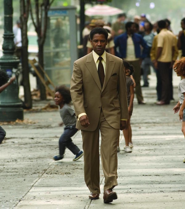 Cut generously, Lucas wears a sand-drift brown, single-breasted suit with peak lapels. A look that isn’t overtly gangster nor overtly businessman. He matches his pocket square with his tie, worn with a spear-pointed collar shirt with brown leather medallion oxford shoes.