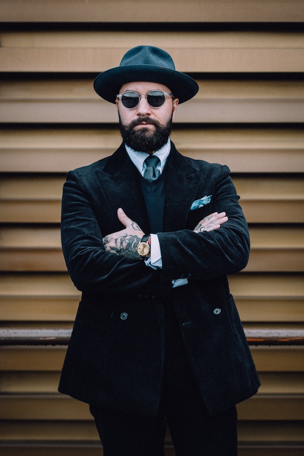 Jared Acquaro demonstrating the art of tonal simplicity with a peaked lapel, double-breasted black corduroy suit and matching trilby hat.