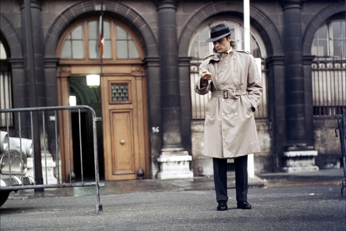 Alain Delon wears a taupe double-breasted trench coat with a belted waist and epaulettes, paired with a grey single-breasted suit and fedora hat in Le Samourai, 1967.