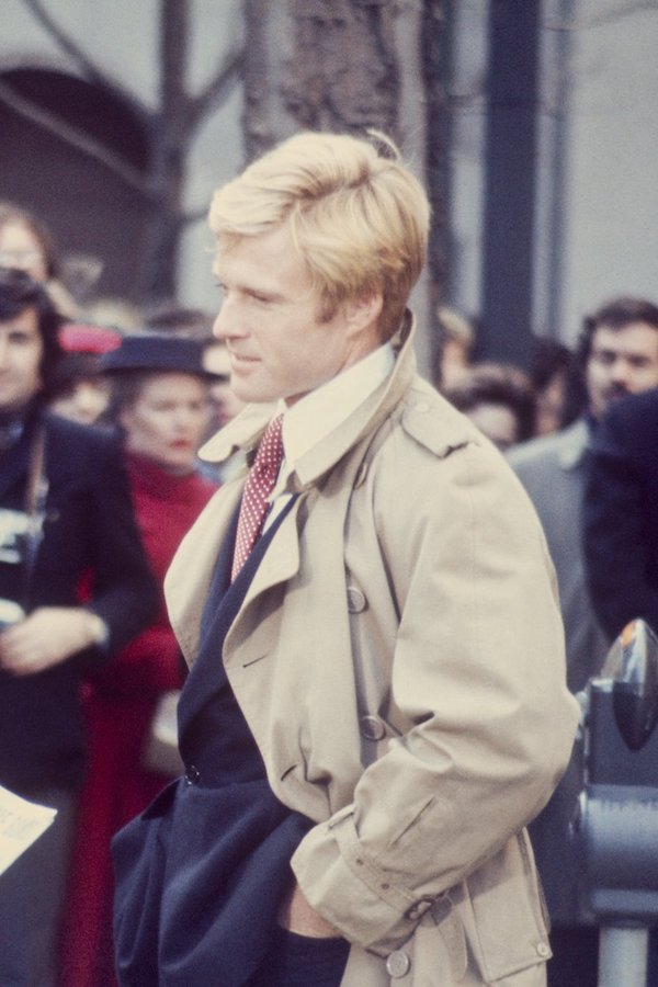 Robert Redford dons a classic taupe trench coat paired with a dark suit and white shirt with a pointed collar. Pictured here in The Way We Were, 1973.