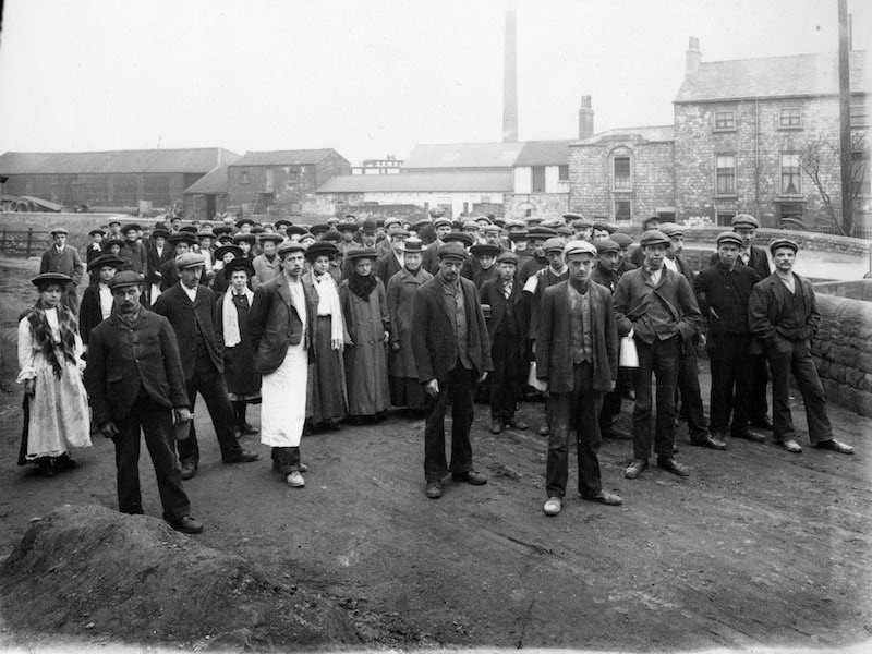 Factory workers sport high-buttoning jackets, mostly single-breasted, and peaked caps in Doncaster, England, circa 1880s.