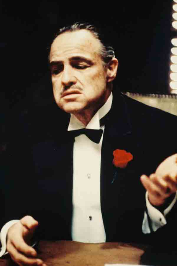 Marlon Brando as Don Vito Corleone wearing a black three-piece dinner suit with silk notched lapels, a white dress shirt, black bow tie and a red floral boutonniere.