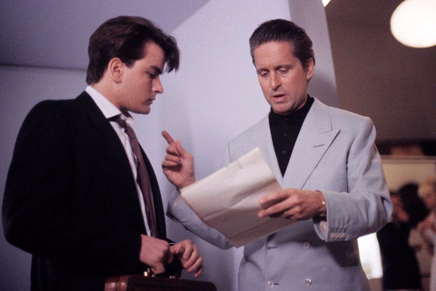 When opting for a more casual look, Gekko wears a light blue 4x2 double-breasted linen jacket with clear buttons over a black polo shirt. Pictured here with Bud Fox played by Charlie Sheen, 1987.