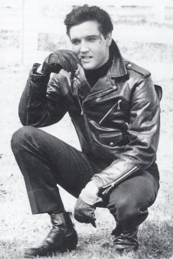 Elvis Presley wears a black leather jacket with a doubled-over front and generous collar which he turns up, paired with driving gloves, leather Chelsea boots and a black sweatshirt in Roustabout, 1964.