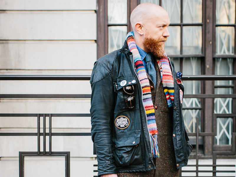 Dean Gomilsek-Cole, Turnbull & Asser's Head of Design & Product Development, dons a vintage wax motorcycle jacket adorned with metal and sewn badges over a tweed Siren Suit, blue denim shirt, tie and hand-knitted scarf. Photograph by Jamie Ferguson.