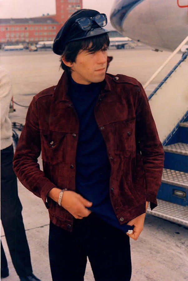 Richards boards a private jet donning a dark tan suede jacket, a navy roll neck and black jeans, topped with peaked cap and Ray Ban sunglasses, 1965.