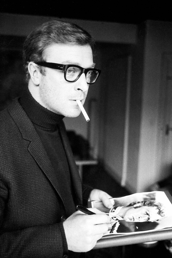 Michael Caine was renowned for his minimalistic tonal style, often pairing a dark roll neck jumper with a similar coloured tailored jacket and his signature dark eye frames. Pictured here signing an autograph, 1966.