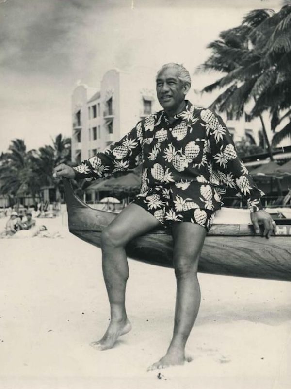 Surfing legend Duke Kahanamoku wears a long sleeve pineapple motif Hawaiian shirt with matching shorts on a beach in Hawaii, circa 1945.