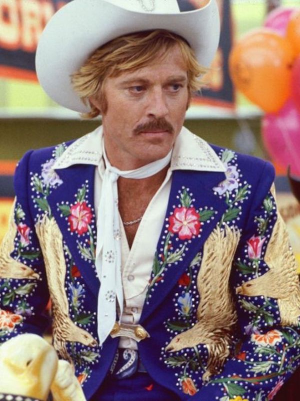 Robert Redford wears a brightly coloured Western-style jacket embroidered with floral motifs and eagles with a white neck scarf and white cowboy hat in The Electric Horseman, 1979.