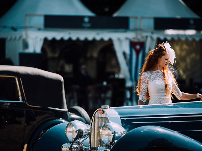 A woman in a lace dress examines a vintage Bugatti.