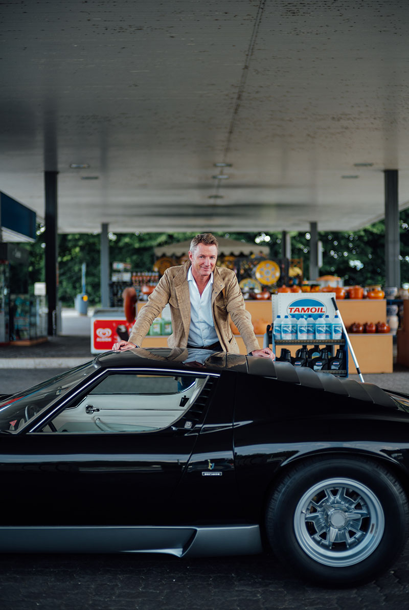 Simon wears a brown jacket with a blue shirt whilst filling up a classic glossy black car.