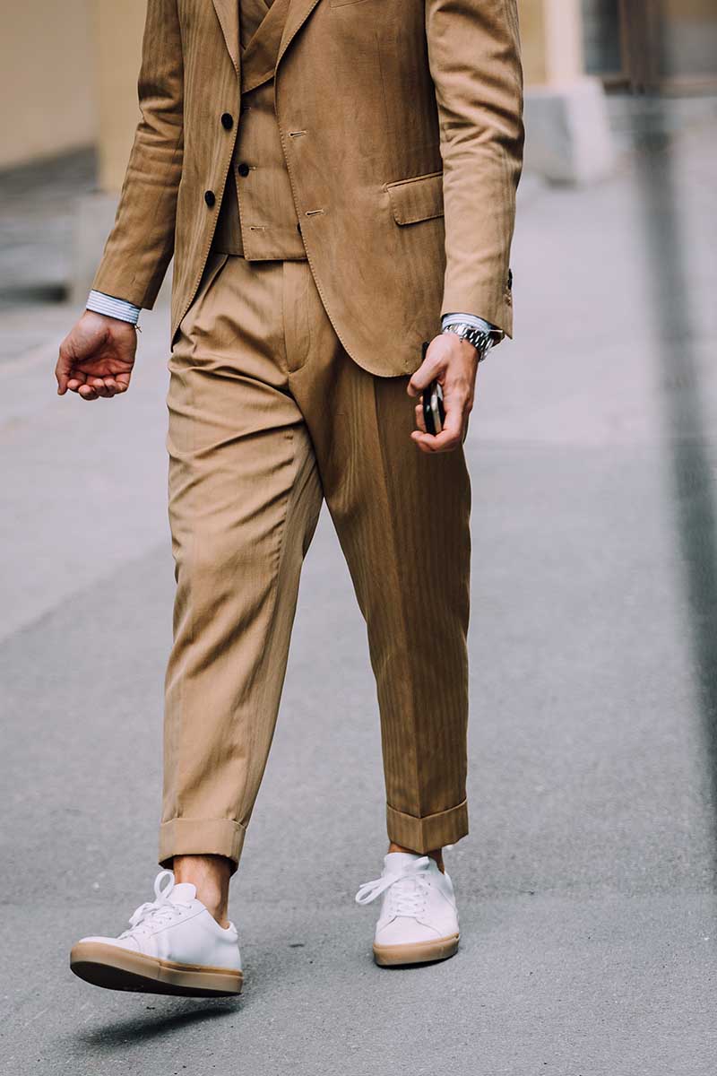 A camel brown three-piece suit styled with crisp white rubbed-soled sneakers. The simple touch of turning up the trouser leg injects excitement into the look.