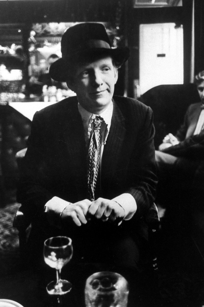 Christopher Gibbs sitting with a drink, wearing a white lace shirt.