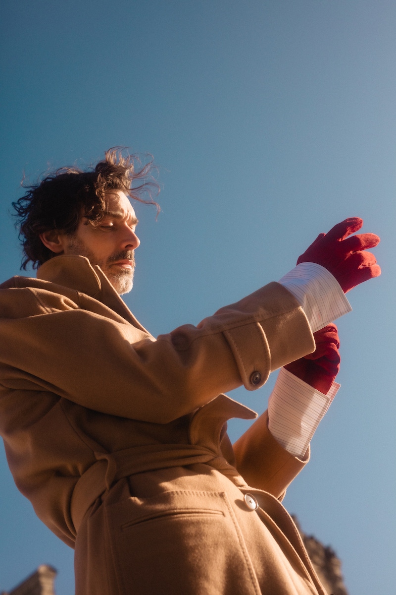 Richard wears: Camel wool Raglan coat, white, red and blue cotton striped pin collar shirt, both Edward Sexton at The Rake; knitted Fair Isle car vest, Connolly; red soft suede leather gloves, Anderson & Sheppard; denim, loafers and tie, models own.